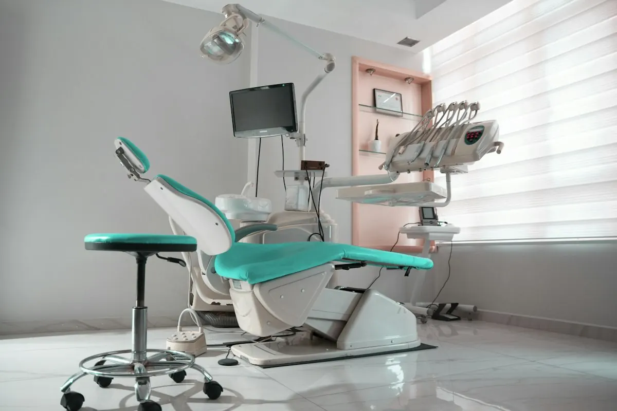 An empty dental treatment room with a teal chair, overhead light and monitor